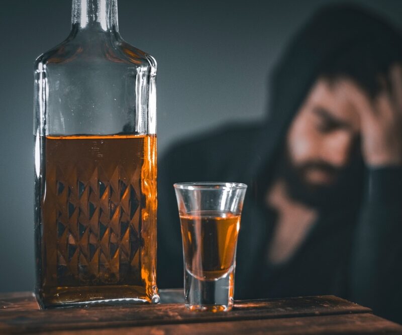 Large bottle of alcohol and a glass filled with alcohol with a man behind wearing a hoodie