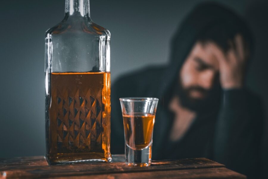 Large bottle of alcohol and a glass filled with alcohol with a man behind wearing a hoodie